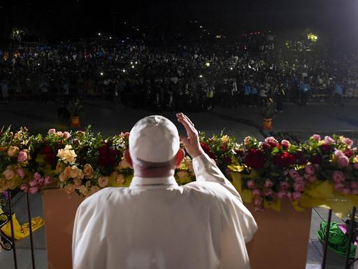 Papst Franziskus steht in Papua-Neuguinea auf einer beleuchteten Bühne vor einer Menschenmenge im Dunkeln, die Lichter in die Höhe hält. 