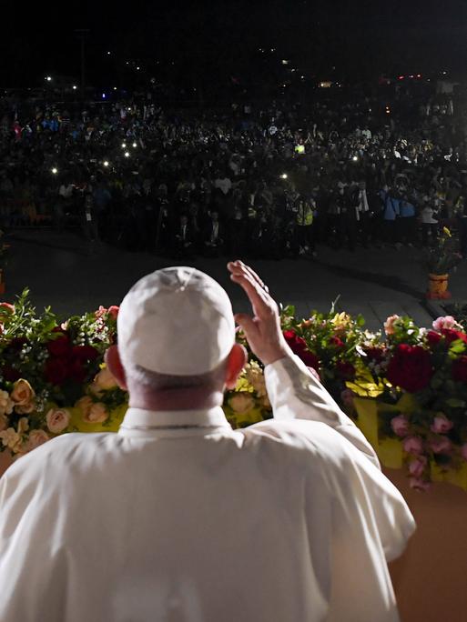 Papst Franziskus steht in Papua-Neuguinea auf einer beleuchteten Bühne vor einer Menschenmenge im Dunkeln, die Lichter in die Höhe hält. 