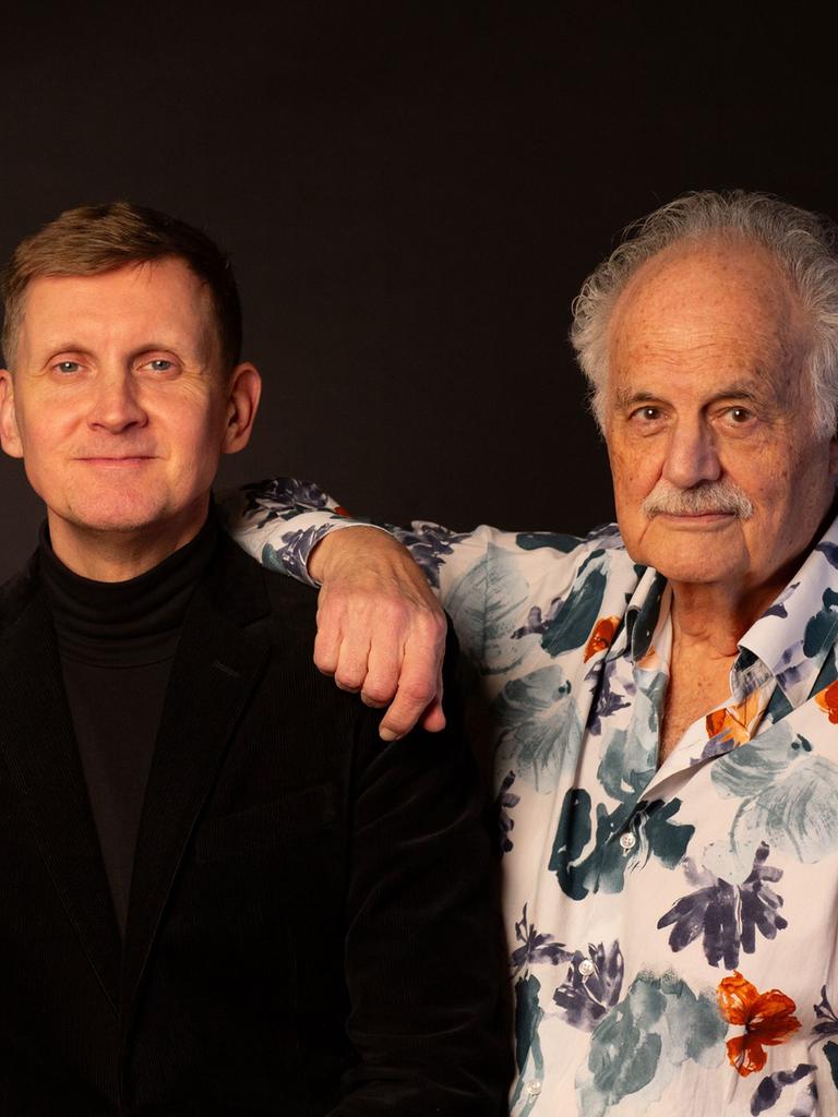 Zwei Männer, einer in dunkler Kleidung, ein anderer mit hellem Hemd mit Blumenmotiven, stehen dicht beieinander und schauen freundlich in die Kamera.