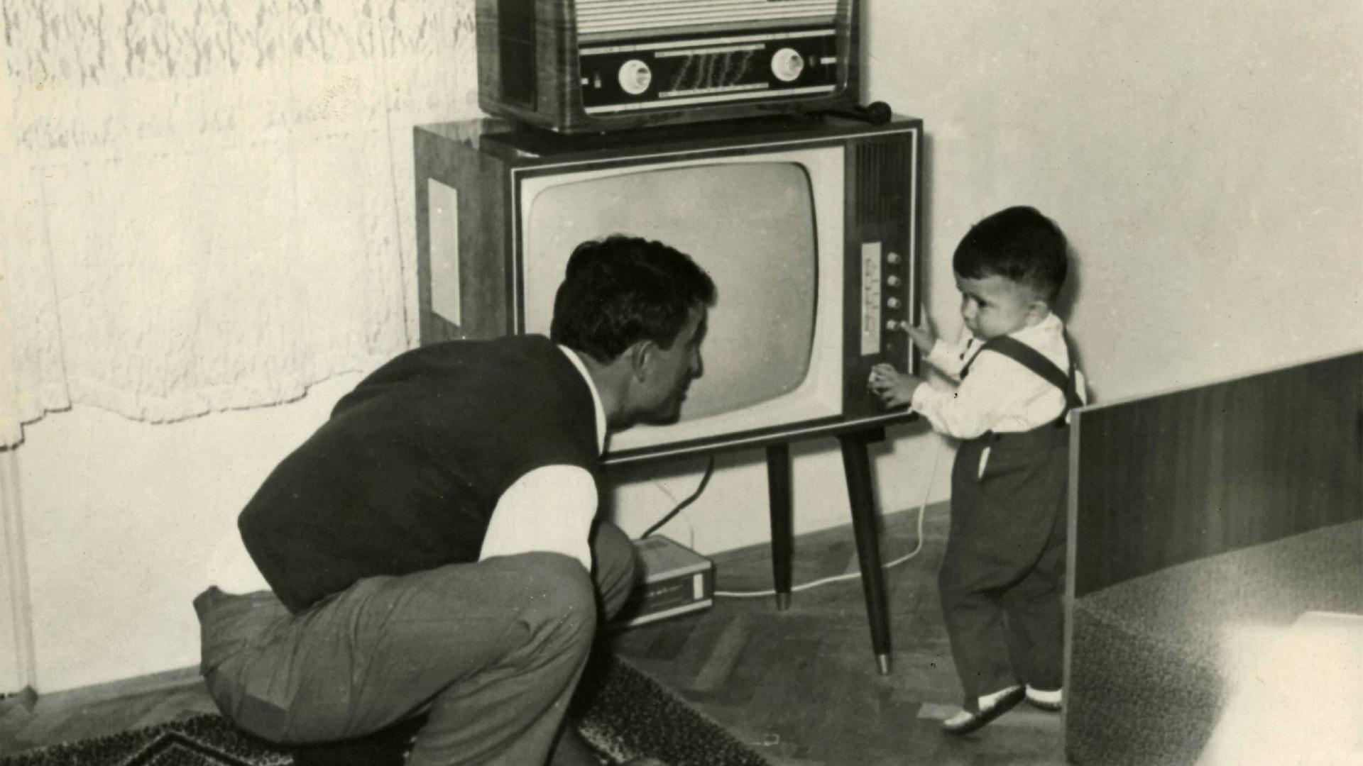 Ein Kleinkind schaltet ca.1950 einen Fernseher ein, darauf steht ein Radio. Der Vater hockt daneben und blickt zu dem Kind. Ein Kleinkind schaltet ca.1950 einen Fernseher ein, darauf steht ein Radio. Der Vater hockt daneben und blickt zu dem Kind.