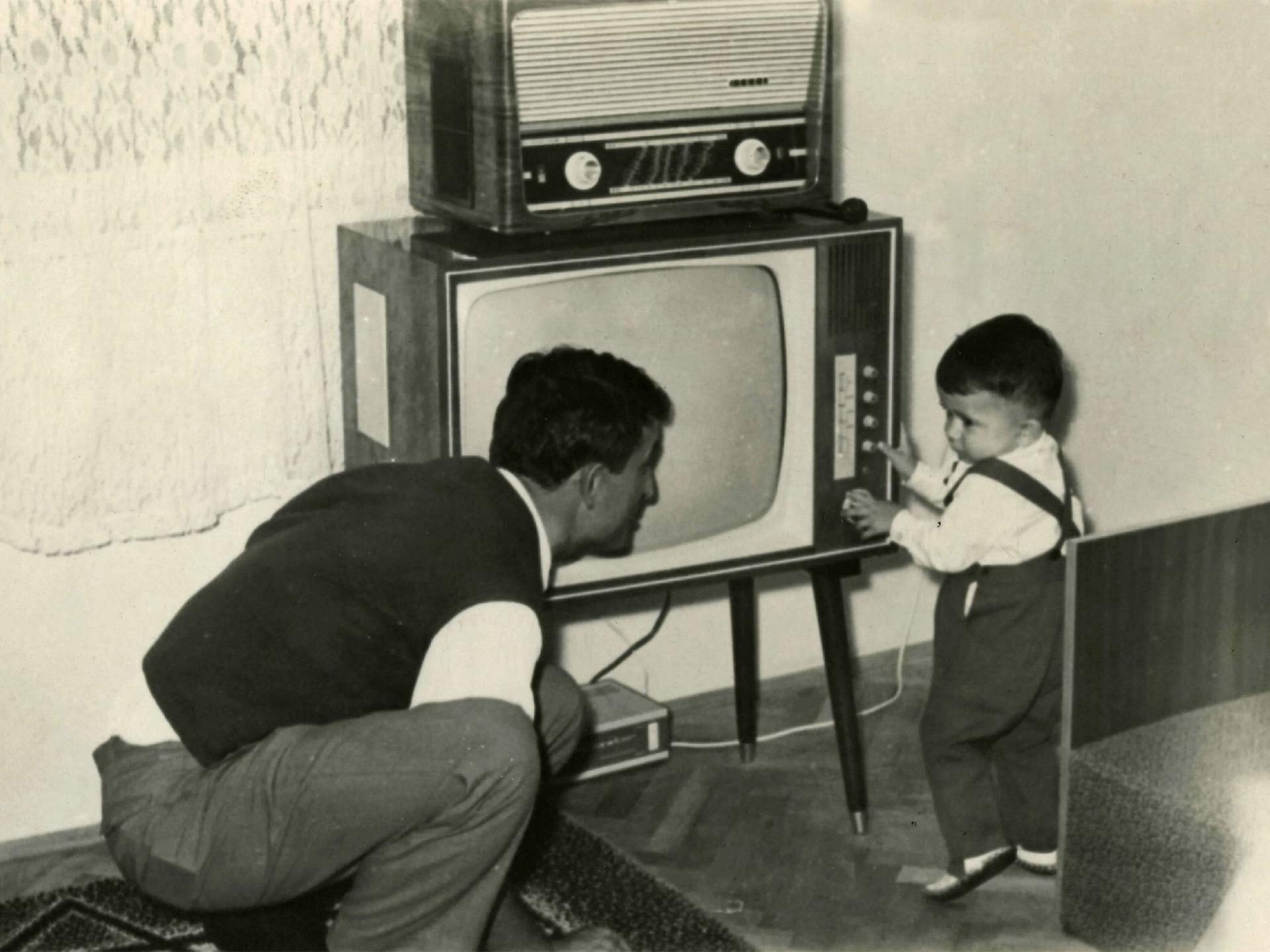 Ein Kleinkind schaltet ca.1950 einen Fernseher ein, darauf steht ein Radio. Der Vater hockt daneben und blickt zu dem Kind.