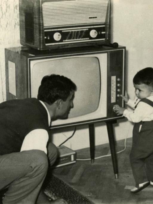 Ein Kleinkind schaltet ca.1950 einen Fernseher ein, darauf steht ein Radio. Der Vater hockt daneben und blickt zu dem Kind.