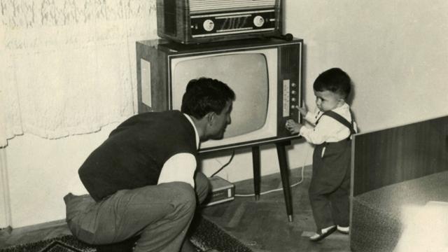 Ein Kleinkind schaltet ca.1950 einen Fernseher ein, darauf steht ein Radio. Der Vater hockt daneben und blickt zu dem Kind.