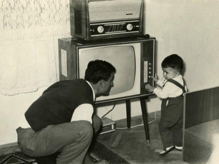 Ein Kleinkind schaltet ca.1950 einen Fernseher ein, darauf steht ein Radio. Der Vater hockt daneben und blickt zu dem Kind.