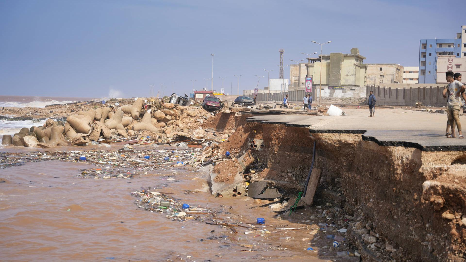 Blick auf die libysche Hafenstadt Derna: Einige Menschen stehen an der Abbruchkante einer Straße, die während der Flutkatastrophe ins Meer gespült wurde. Im Hintergrund sind einige Häuser zu sehen.
