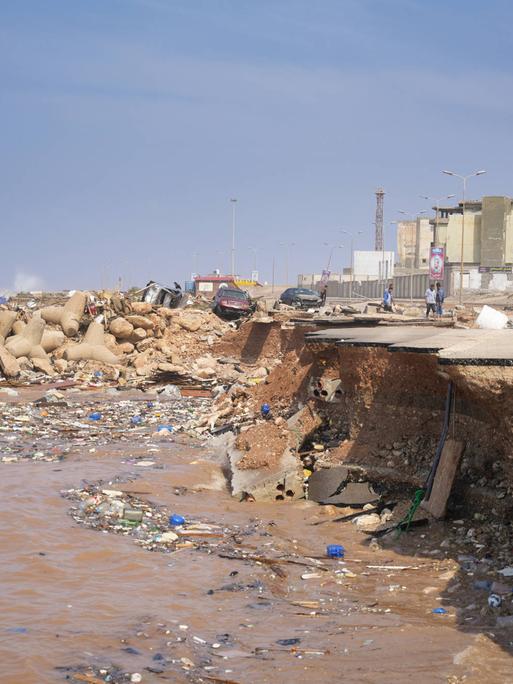 Blick auf die libysche Hafenstadt Derna: Einige Menschen stehen an der Abbruchkante einer Straße, die während der Flutkatastrophe ins Meer gespült wurde. Im Hintergrund sind einige Häuser zu sehen. 