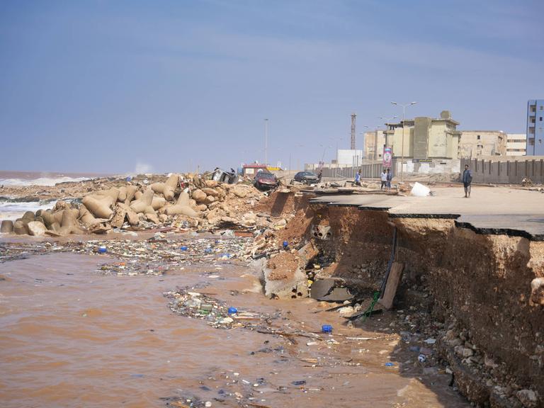 Blick auf die libysche Hafenstadt Derna: Einige Menschen stehen an der Abbruchkante einer Straße, die während der Flutkatastrophe ins Meer gespült wurde. Im Hintergrund sind einige Häuser zu sehen. 