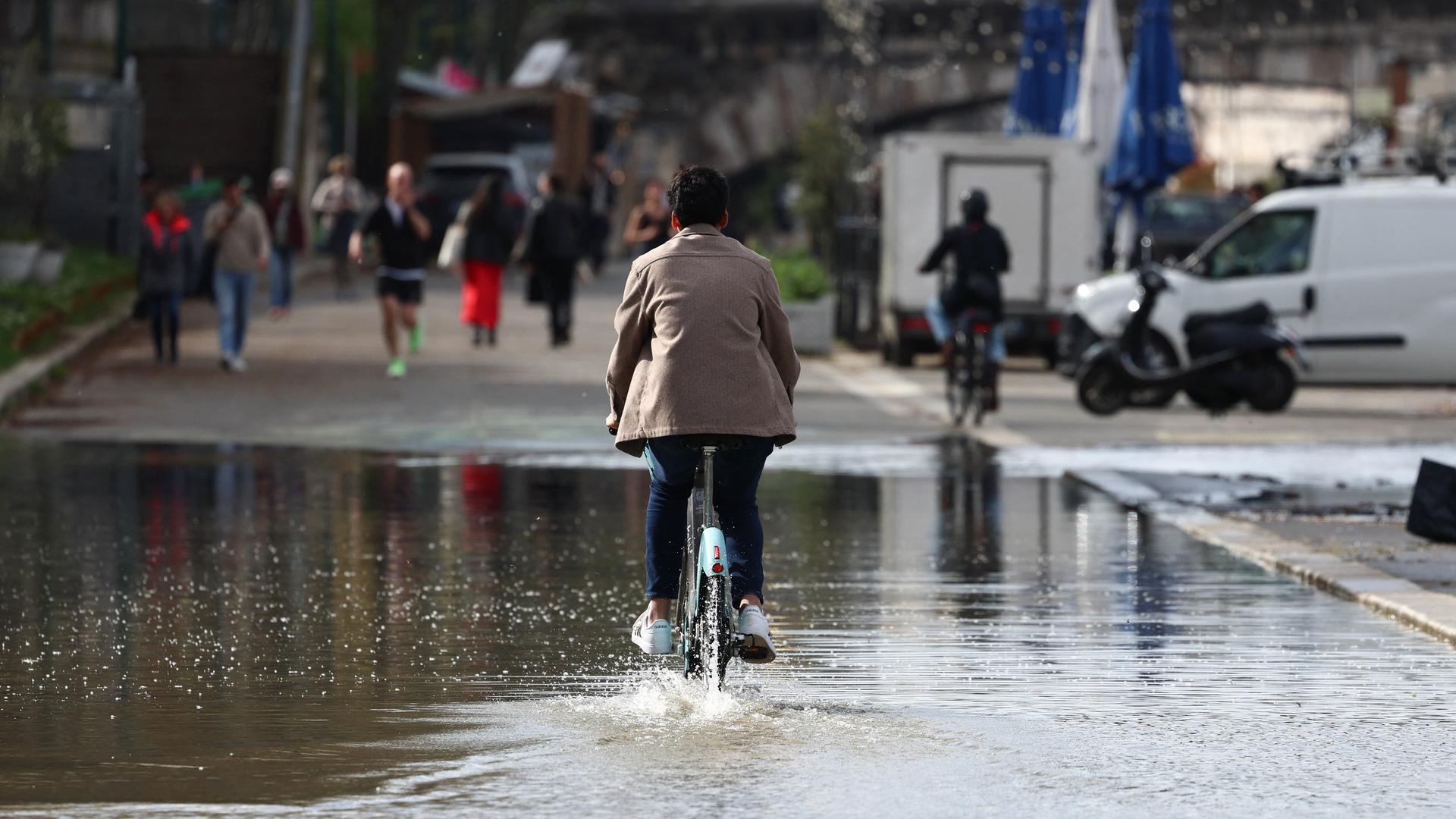 Eine Person auf einem Fahrrad fährt über eine überschwemmte Uferstraße. Im Hintergrund Fußgänger und Jogger. Eine Person auf einem Fahrrad fährt über eine überschwemmte Uferstraße. Im Hintergrund Fußgänger und Jogger.