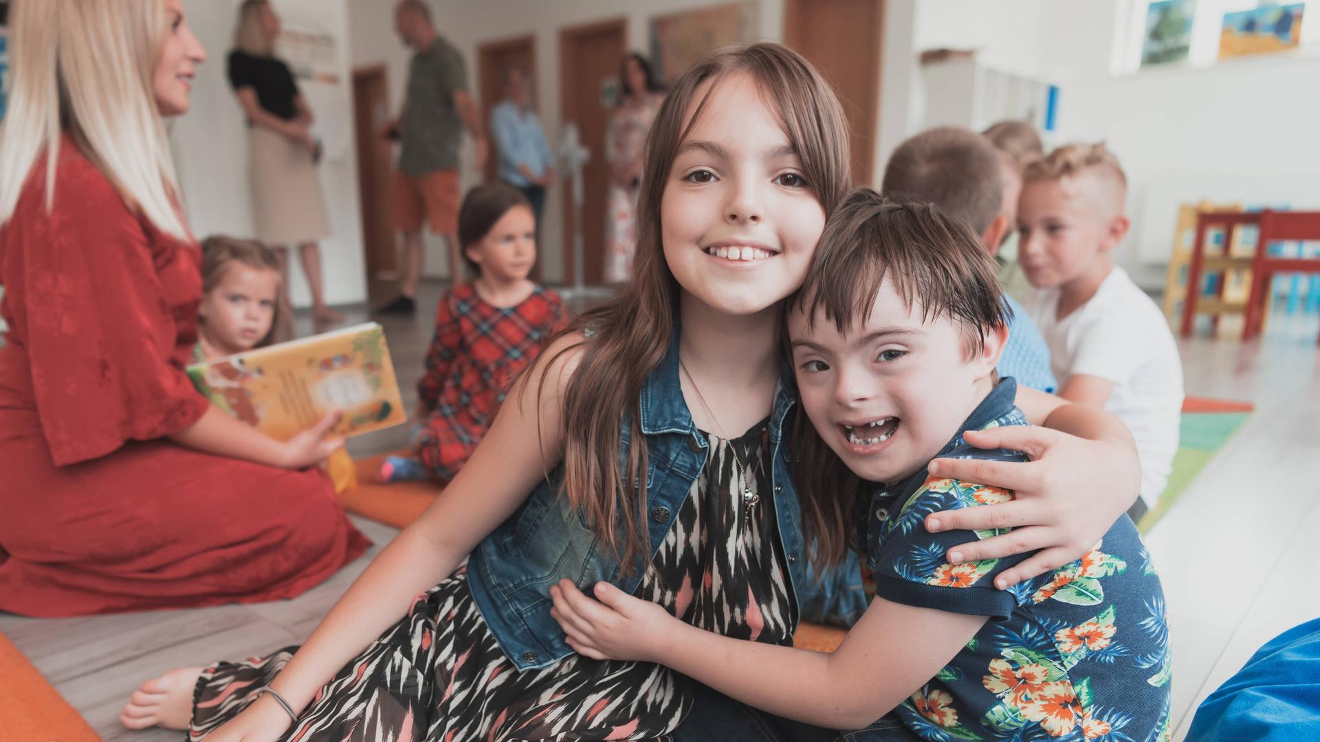 Das Foto zeigt einen lachenden Jungen mit Trisomie-21. Downsyndrom lacht. Er umarmt ein Mädchen. Im Hintergrund sitzen andere Kinder mit einer Betreuerin auf dem Boden.