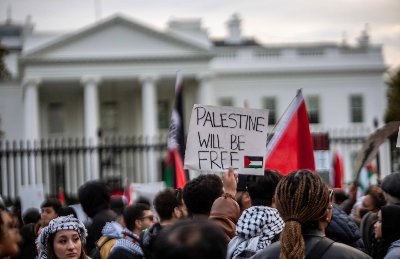 Pro-palästinensische Proteste vor dem Weißen Haus in Washington am 5. November 2023. Das Bild zeigt eine Menschenmenge, die vor einem Zaun steht. Im Hintergrund ist das Weiße Haus leicht verschwommen zu erkennen. Ein Demonstrant hält ein Schild mit der Aufschrift "Palestine will be free" in die Höhe.