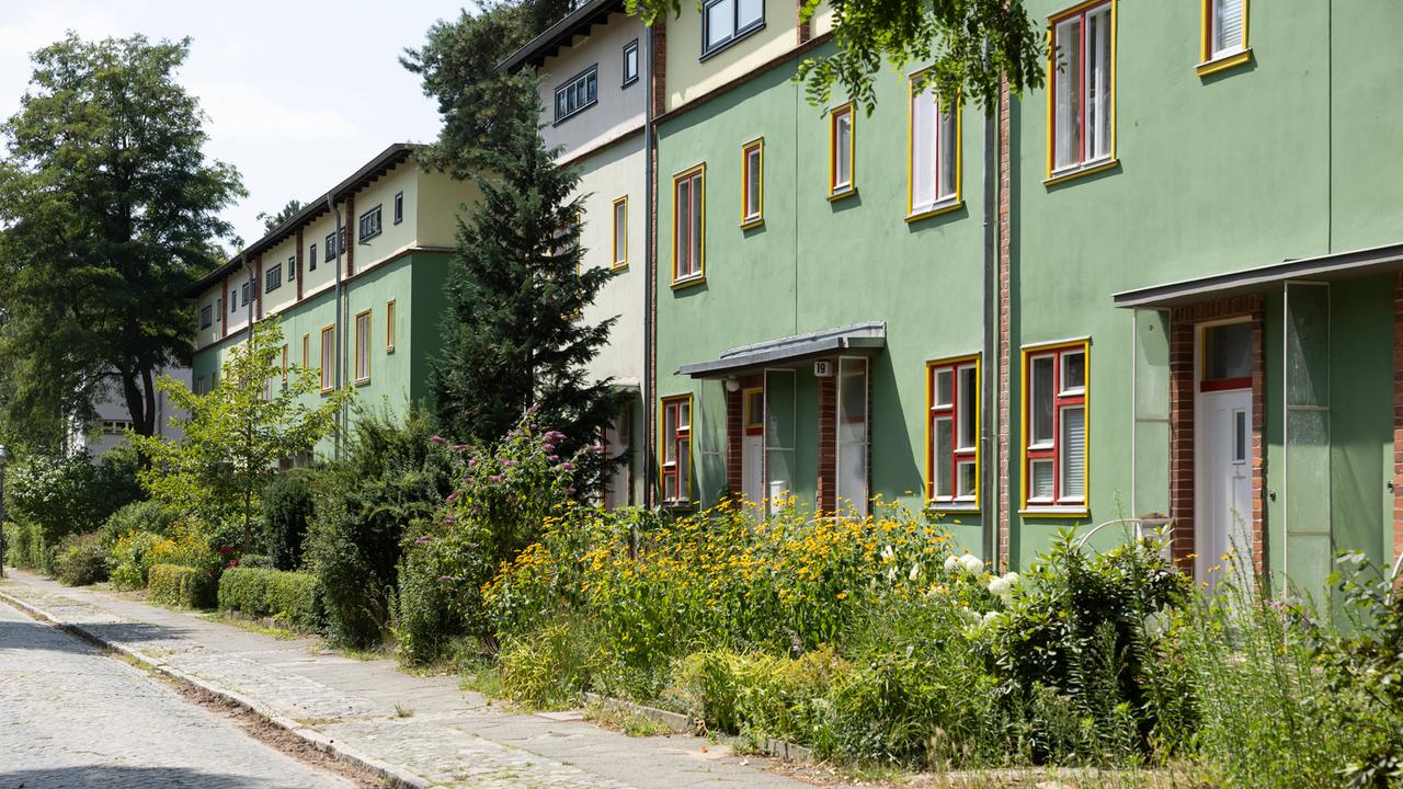 Die Waldsiedlung in Berlin-Zehlendorf: Das Bild zeigt eine Straße mit grünen Gebäuden, bunten Fenstern, Bäumen und Blumen.