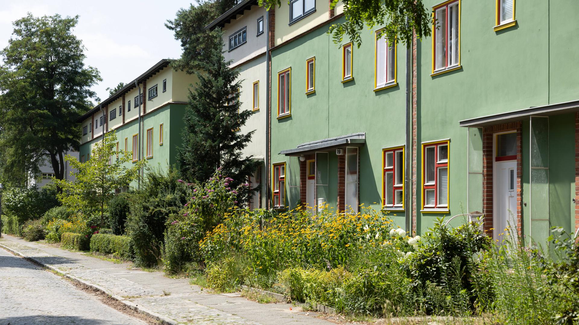 Die Waldsiedlung in Berlin-Zehlendorf: Das Bild zeigt eine Straße mit grünen Gebäuden, bunten Fenstern, Bäumen und Blumen.