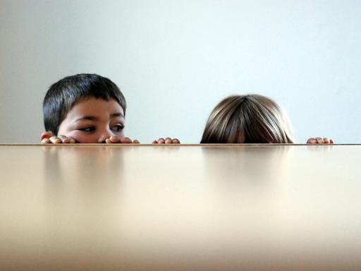 Zwei Kinder blicken über eine Tischplatte. Links ein Junge, von dem das Gesicht fast ganz zu sehen ist; rechts ist lediglich ein Haarschopf erkennbar.