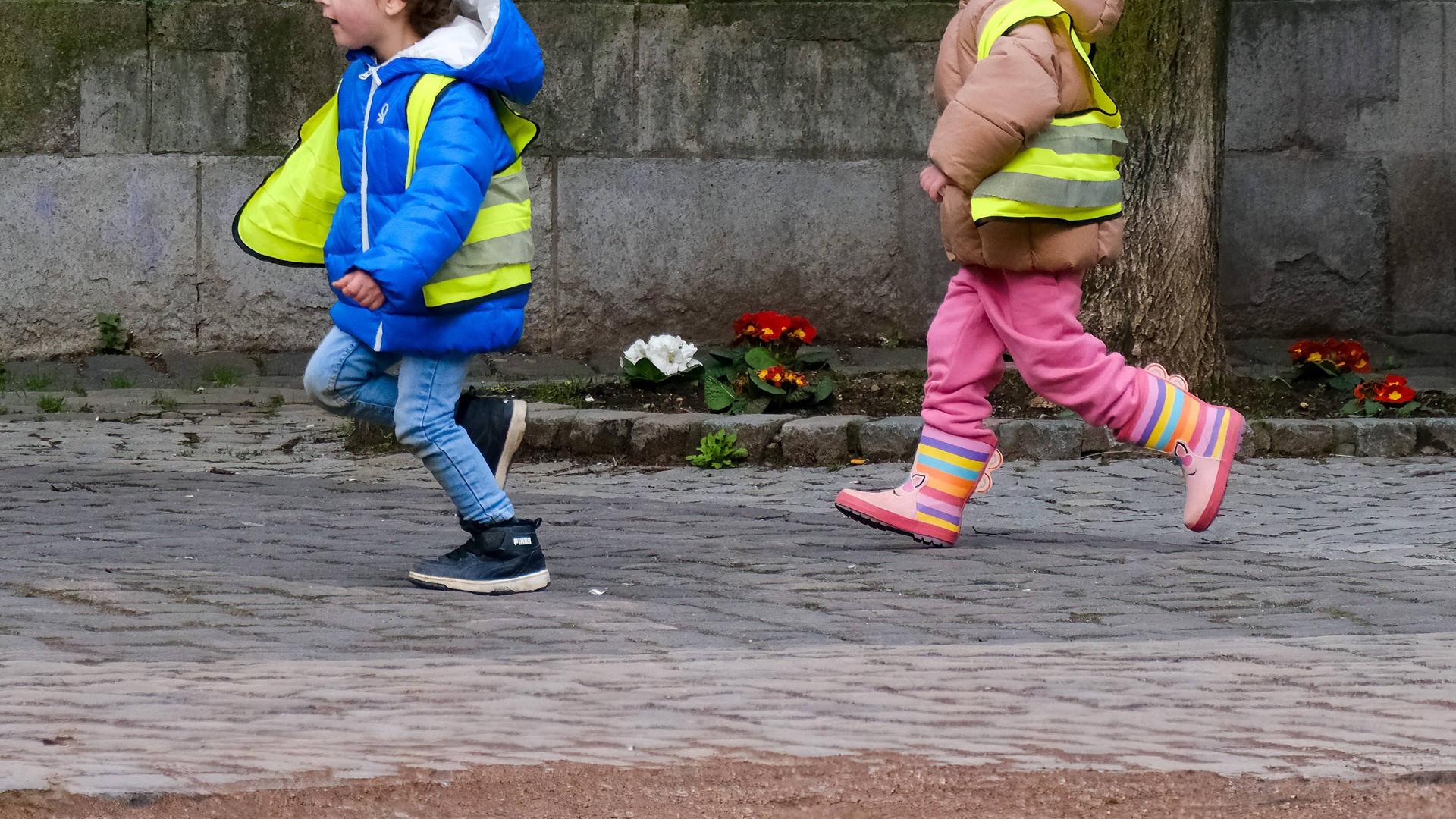Ein Mädchen und ein Junge laufen in Regenkleidung und Warnwesten auf einem Gehweg. 
