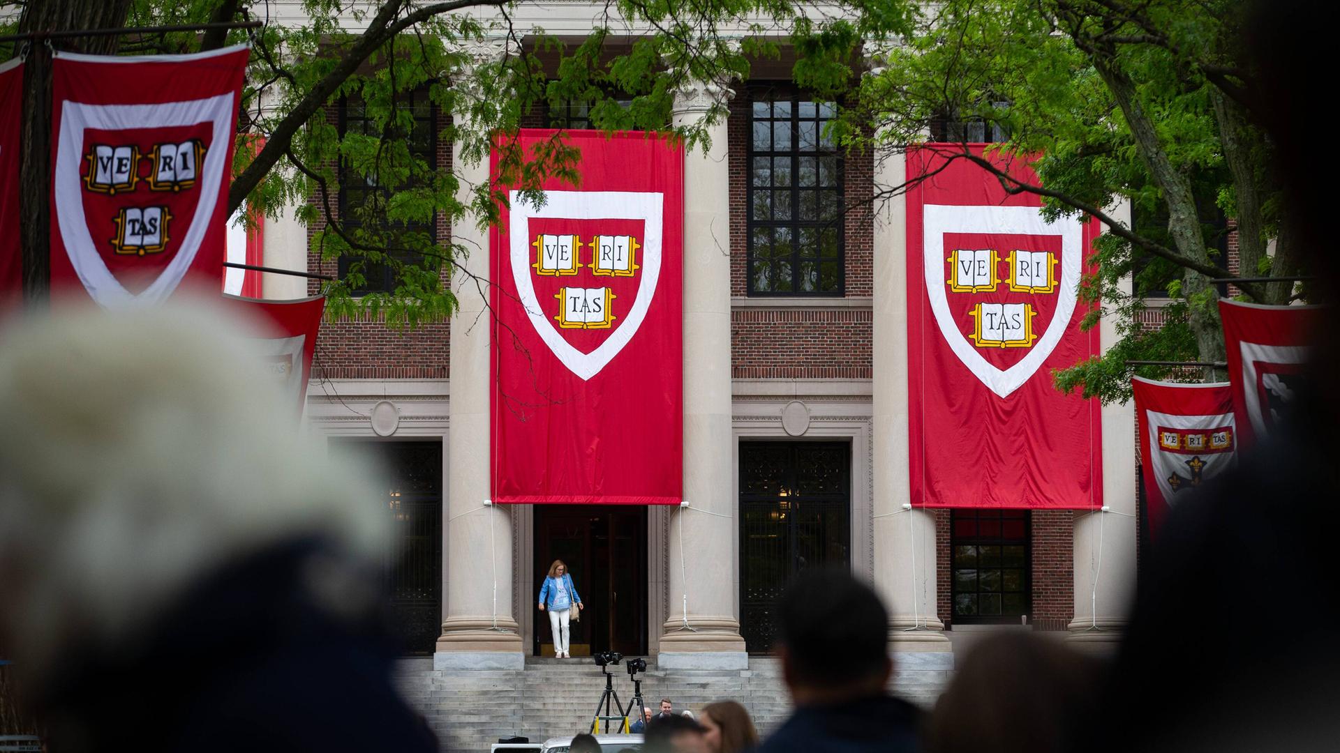 Absolventen, Freunde und Familie versammeln sich an der US-Universität Harvard zur Abschlussrede. Im Vordergrund sind unscharf die Köpfe einiger Menschen zu sehen, hinten Backstein-Uni-Gebäude, an denen Banner mit dem Harvard-Logo hängen. 