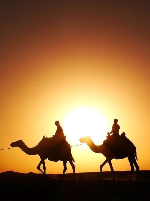 Silhouetten von zwei Reitern auf Kamelen in der Wüste vor einem orangefarbenen Sonnenuntergang.