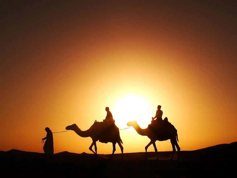 Silhouetten von zwei Reitern auf Kamelen in der Wüste vor einem orangefarbenen Sonnenuntergang.