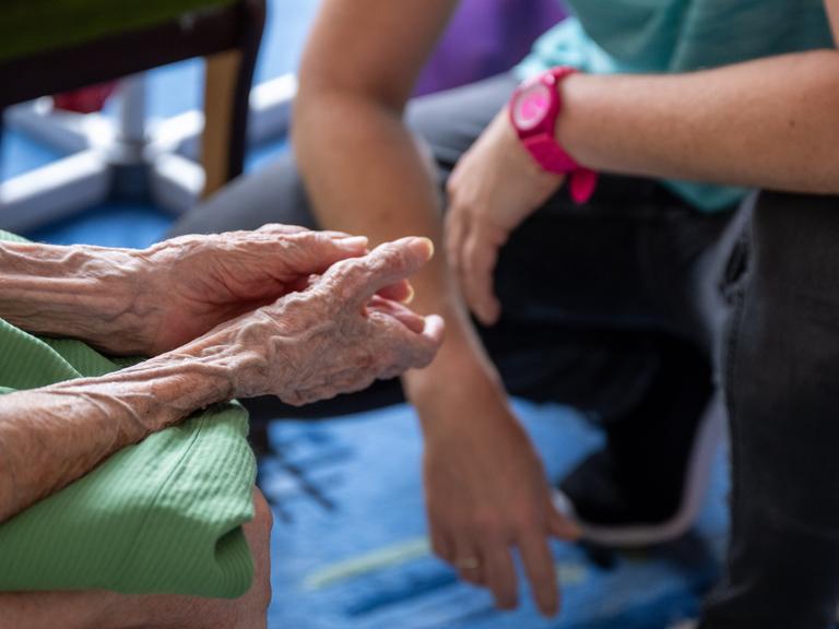 Das Bild zeigt nur die Arme der beiden: Die dünnen faltigen der Patientin auf ihren Knien im Vordergrund; im Hintergrund unscharf die Arme der vor ihr hockenden Pflegerin.
