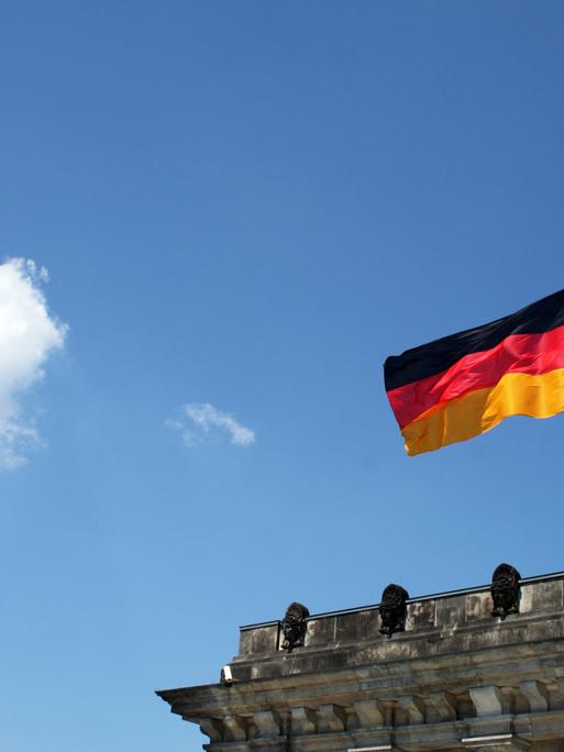 Der Bundestag im ehemaligen Reichstagsgebäude, darüber weht eine Deutschlandfahne, der blaue Himmel öffnet sich und eine Wolke schwebt daher.