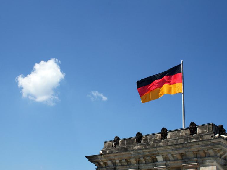 Der Bundestag im ehemaligen Reichstagsgebäude, darüber weht eine Deutschlandfahne, der blaue Himmel öffnet sich und eine Wolke schwebt daher.