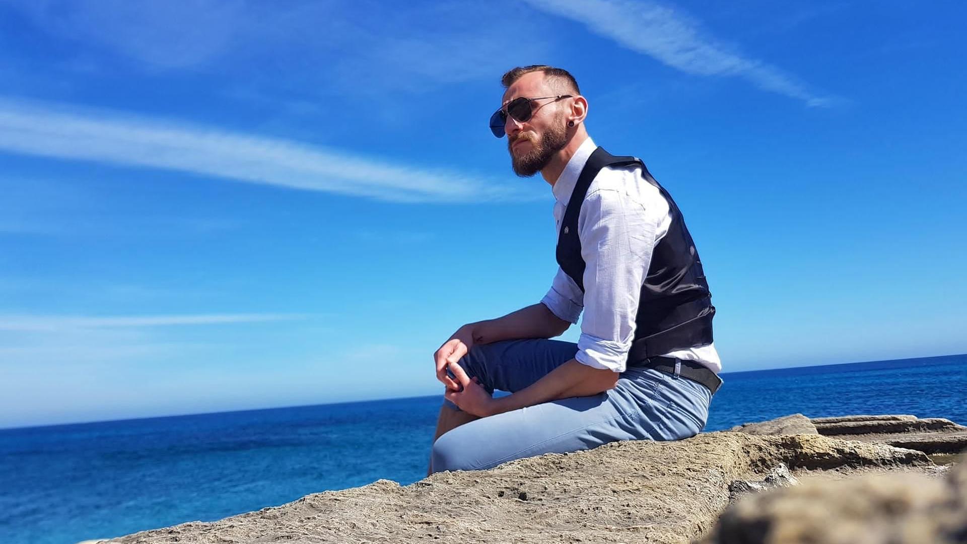 Ein junger Mann mit Sonnenbrille sitzt auf einem Felsen am Meer.