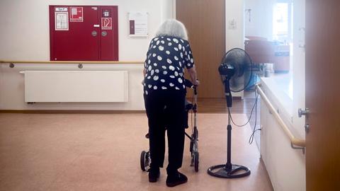 Eine pflegebedürftige Frau in einem Altenheim steht neben einem Ventilator.