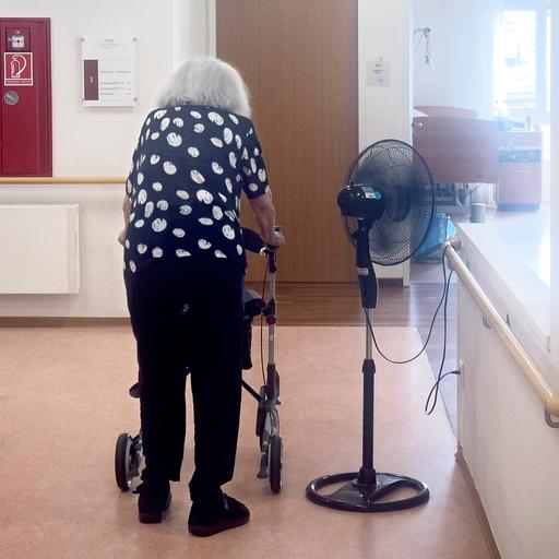 Eine pflegebedürftige Frau in einem Altenheim steht neben einem Ventilator.