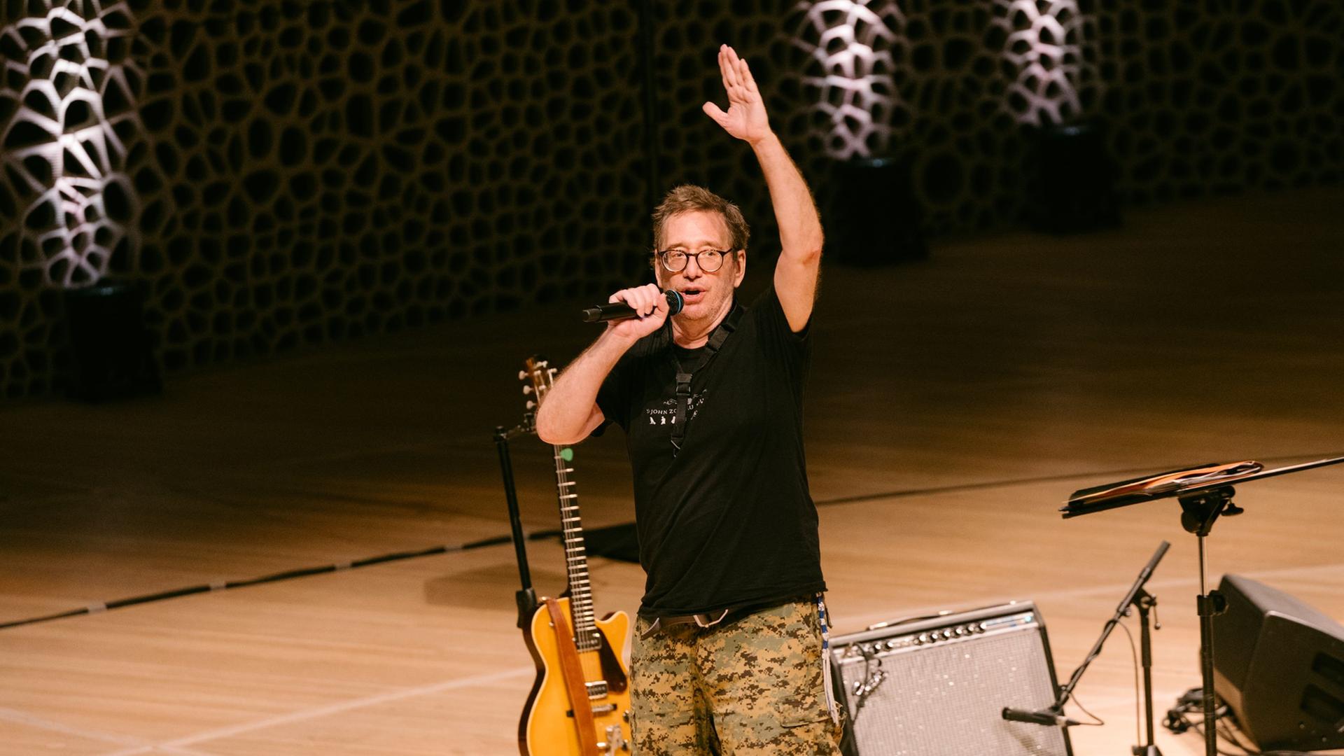 Ein Mann mit Brille  und lockerer Kleidung steht auf einer weiten Bühne, spricht in eine Mikrofon und hebt dabei eine Hand.