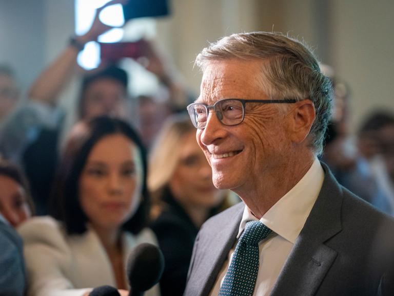 Tech-Millardär Bill Gates bei einer Veranstaltung der Bill & Melinda Gates Foundation. Er trägt eine Brille, einen grauen Anzug und lacht in die Kameras. Im Hintergrund steht eine Reporterin, die ihm ein Mikrofon hinhält. 