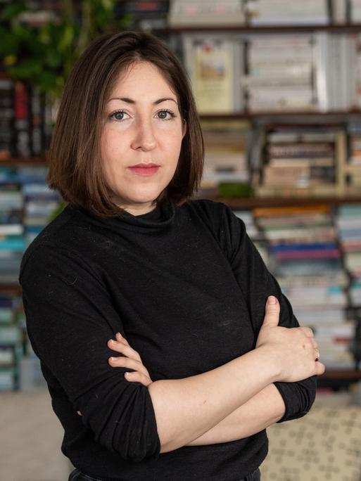 Deborah Feldman steht mit verschränkten Armen in ihrem Wohnzimmer und schaut in die Kamera. Sie hat einen schulterlangen dunkelbraunen Bob und trägt einen schwarzen Rollkragenpulli.