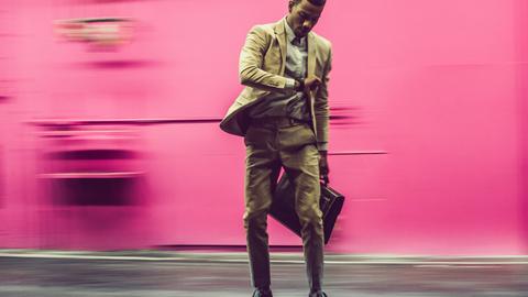 Ein Mann im Businessanzug fährt vor einer rosa Wand auf einem Skateboard. Dabei schaut er auf die Uhr. In der anderen Hand hat er eine Aktentasche.