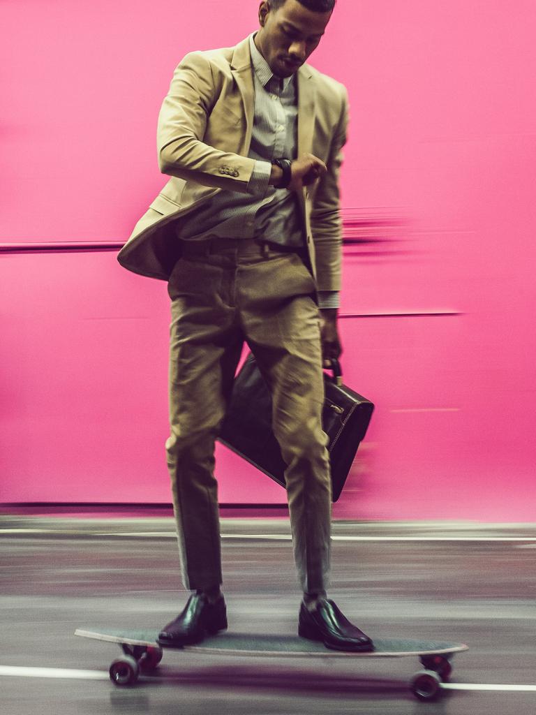 Ein Mann im Businessanzug fährt vor einer rosa Wand auf einem Skateboard. Dabei schaut er auf die Uhr. In der anderen Hand hat er eine Aktentasche.