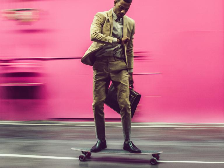Ein Mann im Businessanzug fährt vor einer rosa Wand auf einem Skateboard. Dabei schaut er auf die Uhr. In der anderen Hand hat er eine Aktentasche.