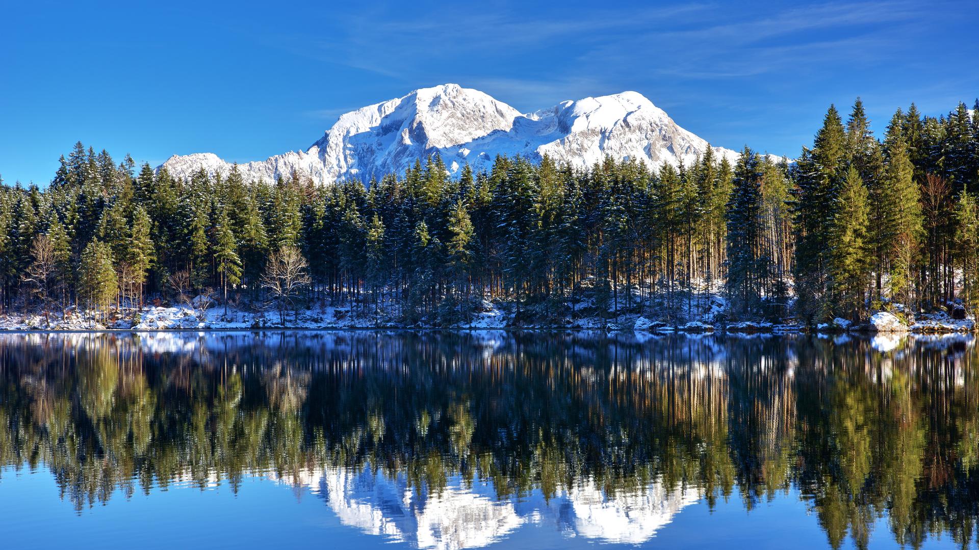 Ein Foto von einem klaren See, in dem sich Bäume spiegeln. Im Hintergrund ist ein Berg mit Schneekuppe zu sehen.