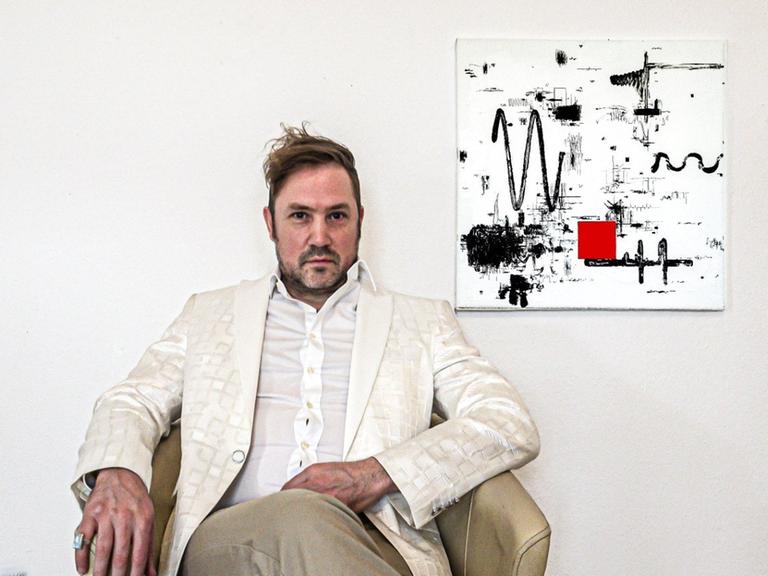 Ein Mann in hellem Seidensakko sitzt in einem Sessel und schaut in die Kamera, während nebem ihm ein abstraktes Kunstwerk in weiß/schwarz/rot an der Wand hängt.