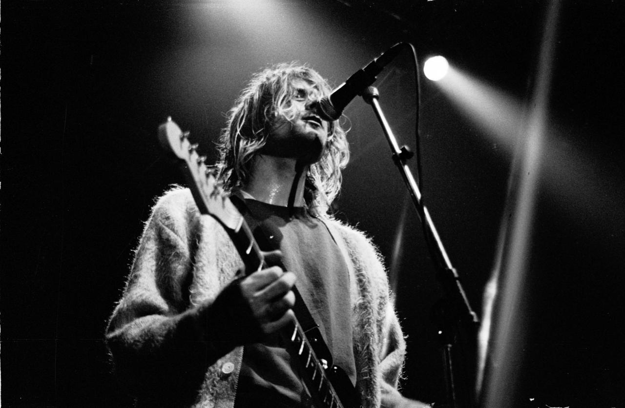 Schwarzweißfoto von Kurt Cobain, der mit seiner Gitarre auf der Bühne steht und in ein Mikrofon singt.