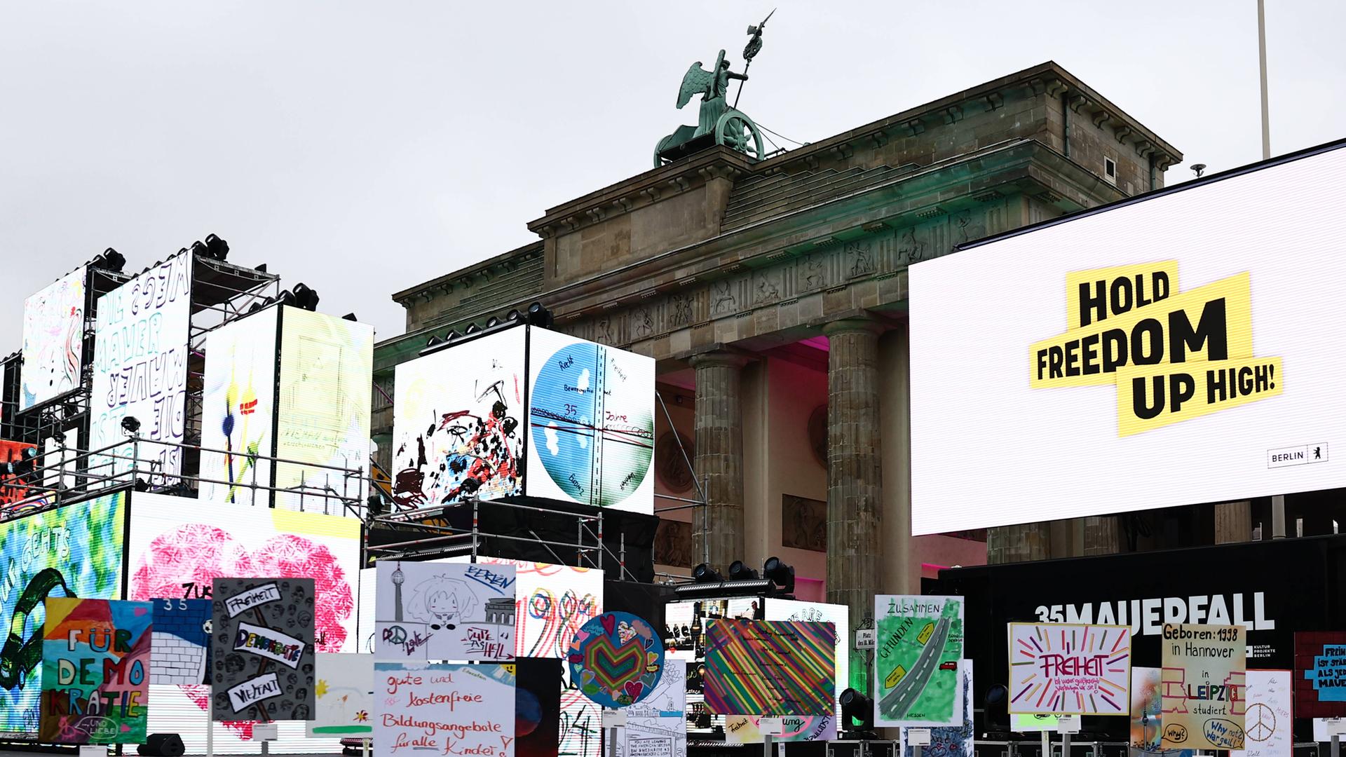 Vor dem Brandenburger Tor sind viele bunte Plakate nebeneinander aufgestellt. Im Hintergrund sind Videoleinwände zu sehen. 