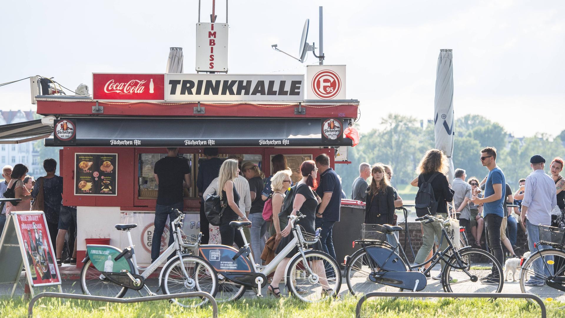 Ein Büdchen am Rheinufer in Düsseldorf, die Menschen stehen Schlange für ein Getränk an einem Sommerabend."Trinkhalle" und "Imbiss" steht auf dem Dach.