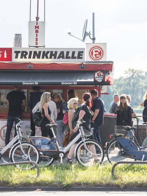 Ein Büdchen am Rheinufer in Düsseldorf, die Menschen stehen Schlange für ein Getränk an einem Sommerabend."Trinkhalle" und "Imbiss" steht auf dem Dach.