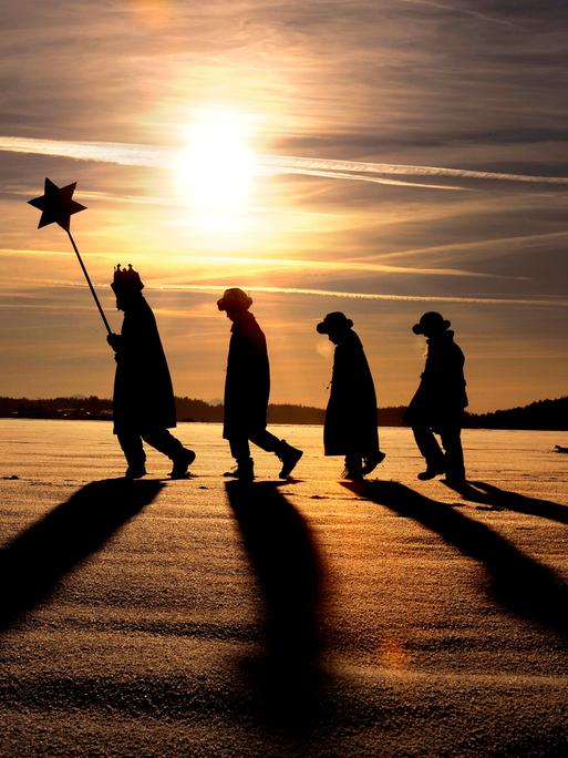 Vier Sternsinger laufen durch die verschneite Landschaft, im tiefstehenden Abendsonnen werfen sie lange Schatten. (Katholische Gemeinde Hl. Familie Kaufbeuren)