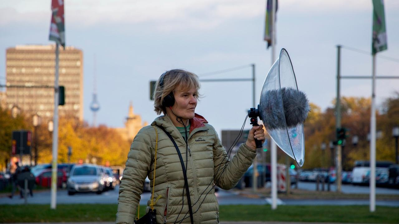 Eine Frau steht auf einem Grünstreifen inmitten des Berliner Autoverkehrs und hat ein Mikrofon mit einem großen Windschutz in der Hand.