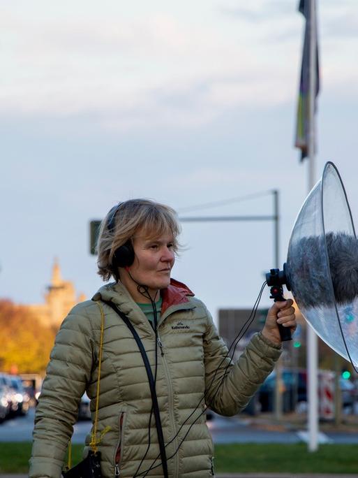 Eine Frau steht auf einem Grünstreifen inmitten des Berliner Autoverkehrs und hat ein Mikrofon mit einem großen Windschutz in der Hand.