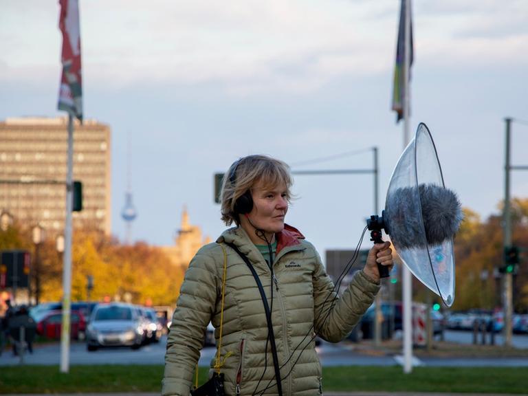 Eine Frau steht auf einem Grünstreifen inmitten des Berliner Autoverkehrs und hat ein Mikrofon mit einem großen Windschutz in der Hand.