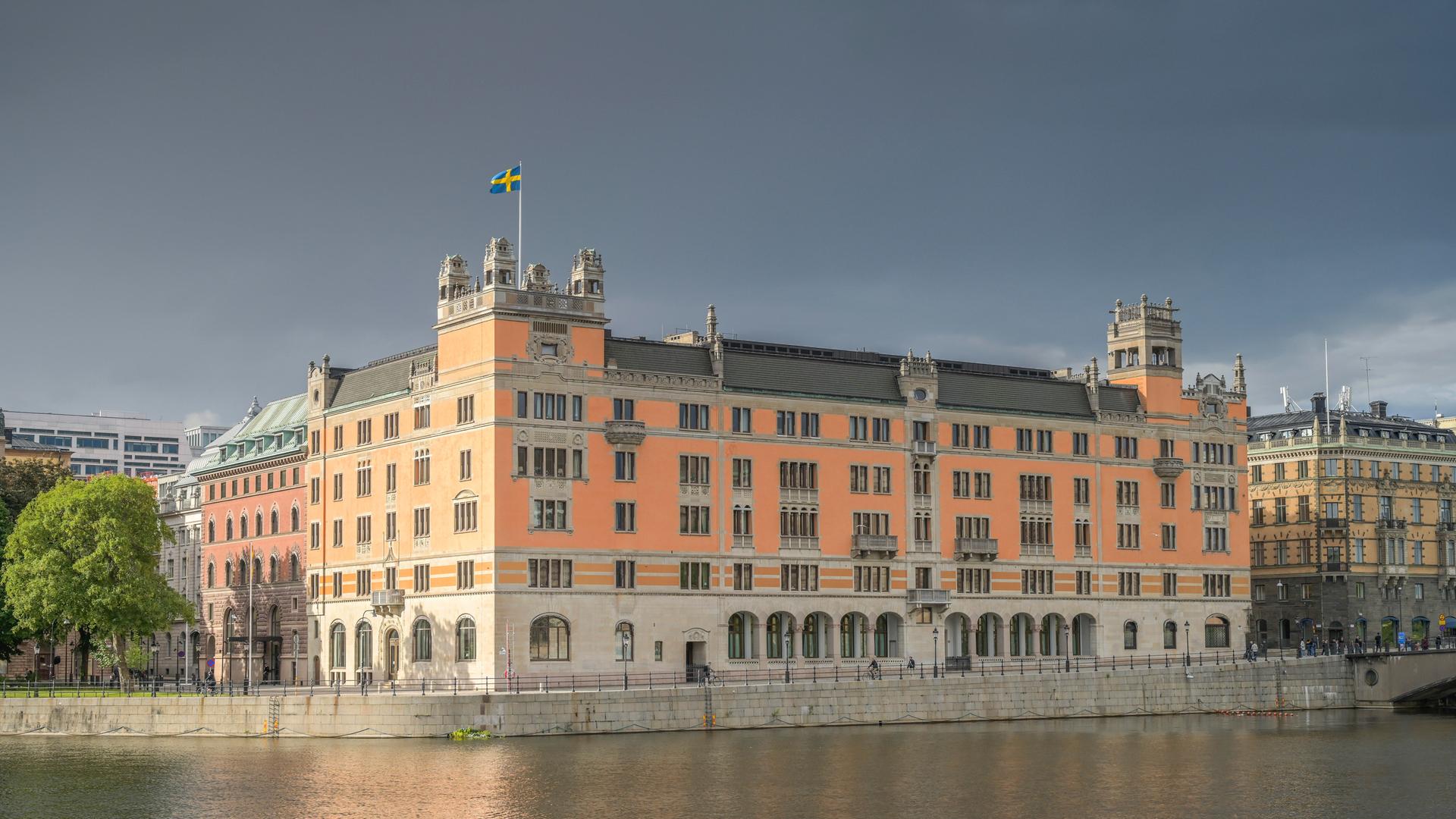 Das große orangefarbene Gebäude aus der Entfernung gesehen. Davor eine Wasserfläche.