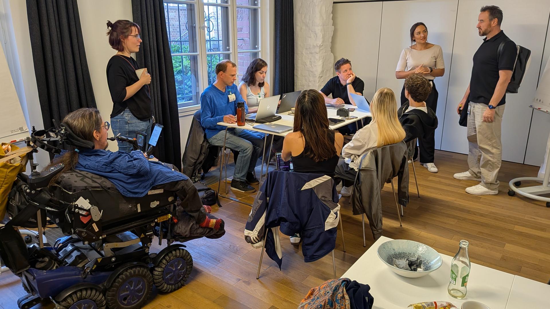 Das Foto zeigt eine Gruppe von Menschen, die an einem Tisch sitzen. Eine Person sitzt in einem besonderen Roll-Stuhl. Das Foto zeigt eine Gruppe von Menschen, die an einem Tisch sitzen. Eine Person sitzt in einem besonderen Roll-Stuhl.