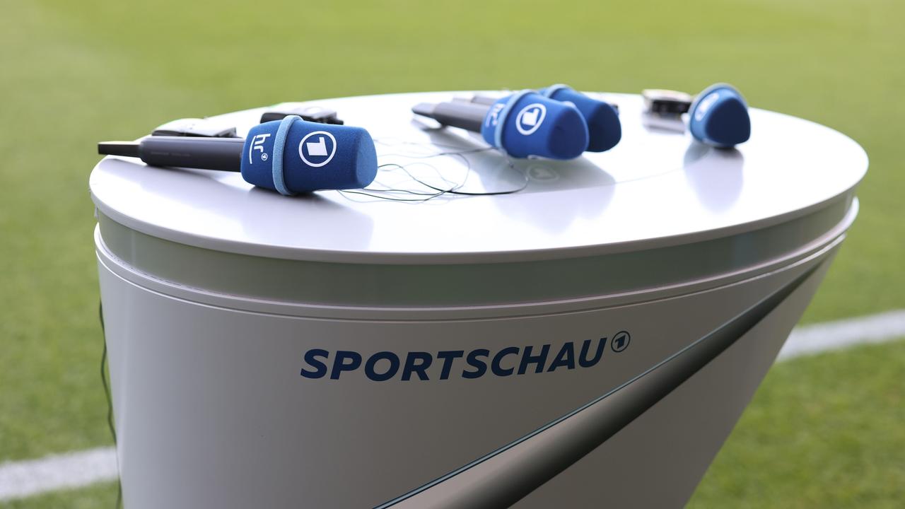 Mehrere ARD-Mikrofone liegen aus einen "Sportschau"-Tisch am Spielfeldrand eines Stadions. 