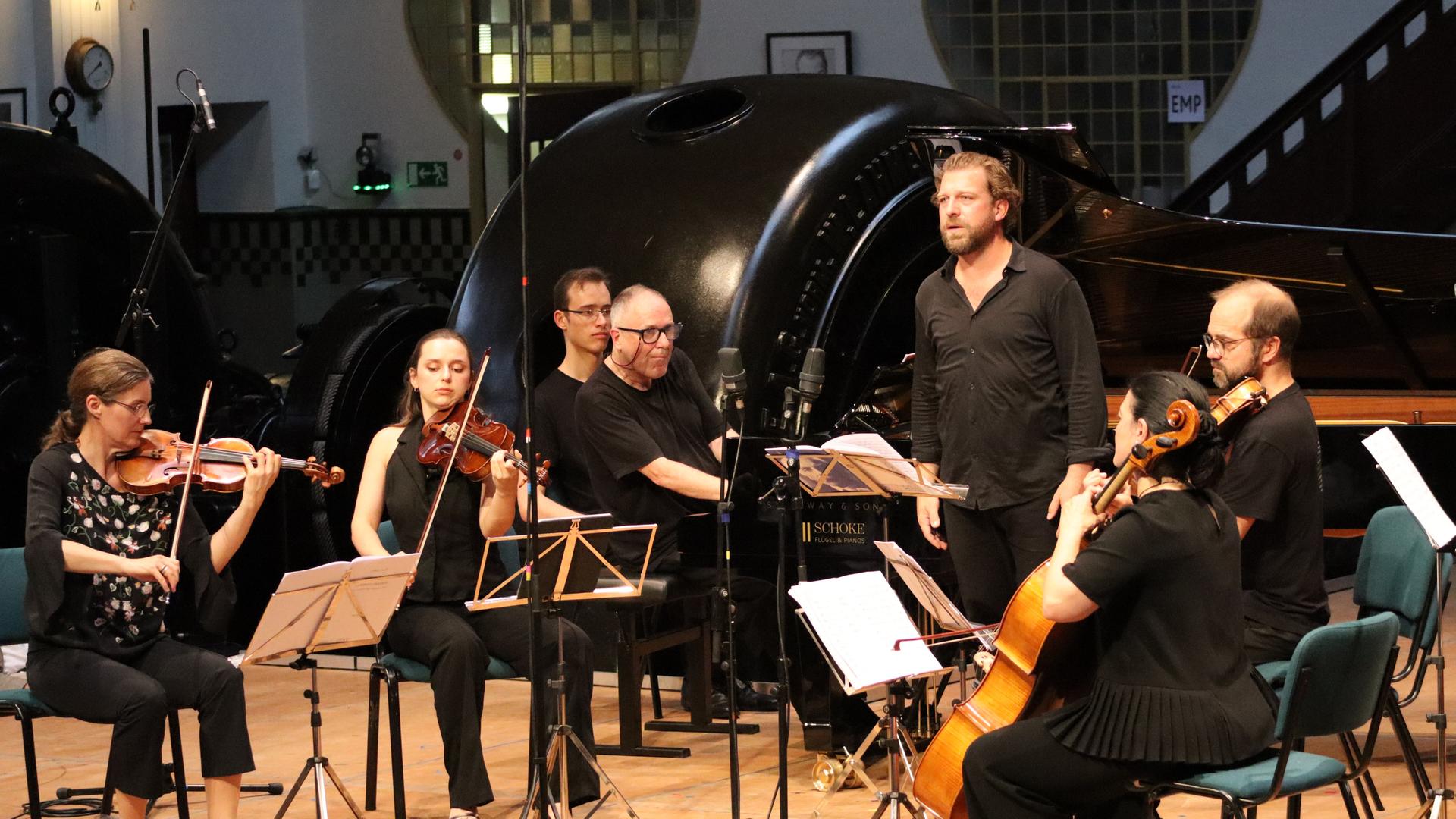 Ein Streichquartett, ein Pianist am Flügel und ein stehender Sänger auf einer Bühne in Konzertsituation. Alle sind schwarz gekleidet, im Hintergrund sieht man den historischen Raum mit einer stillgelegten Turbine. 