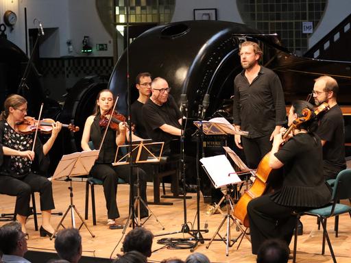 Ein Streichquartett, ein Pianist am Flügel und ein stehender Sänger auf einer Bühne in Konzertsituation. Alle sind schwarz gekleidet, im Hintergrund sieht man den historischen Raum mit einer stillgelegten Turbine. 