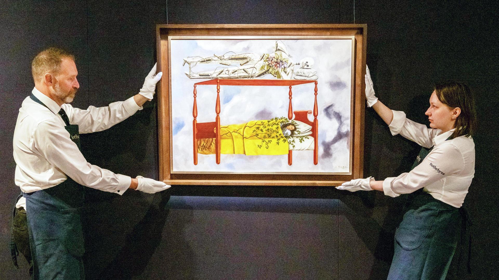 Ein Mann und eine Frau halten ein Gemälde von Frida Kahlo. Es zeigt eine Frau in einem Bett.