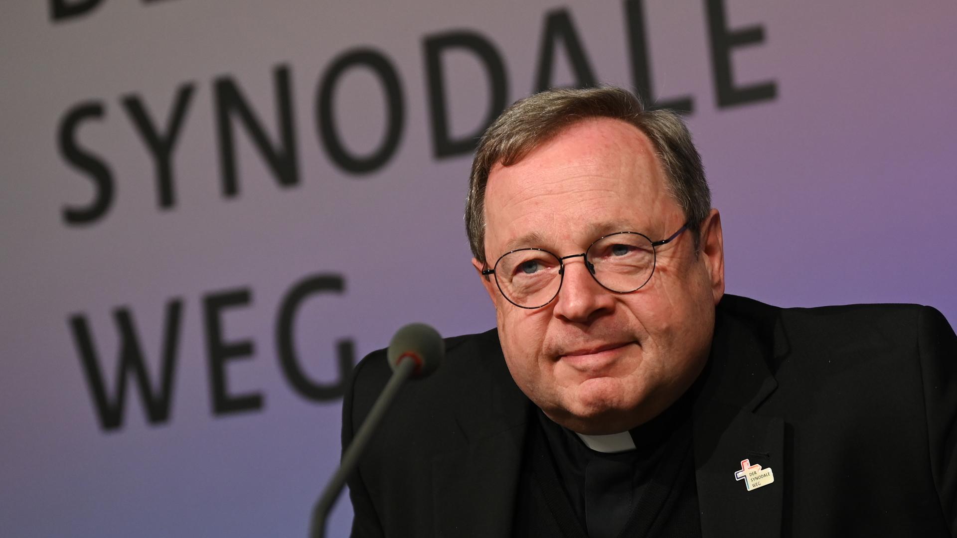 Georg Bätzing, Bischof von Limburg und Vorsitzender der Deutschen Bischofskonferenz, mit Brille und einem Kreuz am Revers, sitzt hinter einem Mikrofon und vor einem Schild, auf dem "Der synodale Weg" steht. Georg Bätzing, Bischof von Limburg und Vorsitzender der Deutschen Bischofskonferenz, mit Brille und einem Kreuz am Revers, sitzt hinter einem Mikrofon und vor einem Schild, auf dem "Der synodale Weg" steht.